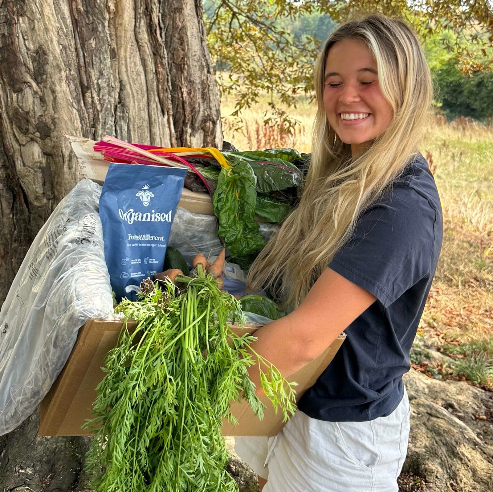 A smiling member holding a box of colorful seasonal veg beneath a tree at My Little Farm