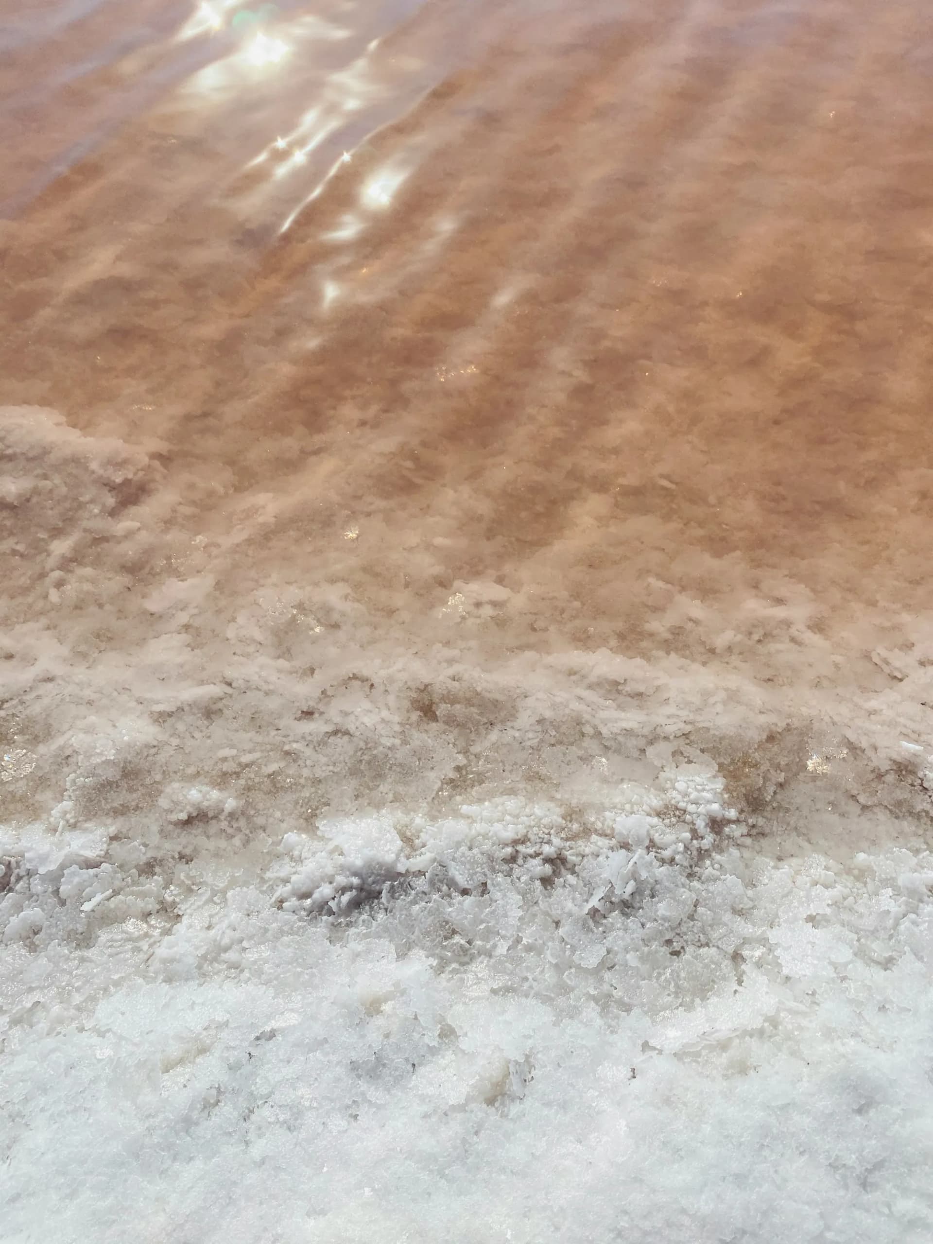 Rust-red brine meets a thick crust of white salt at the edge of a salt flat, sunlight glinting off the water
