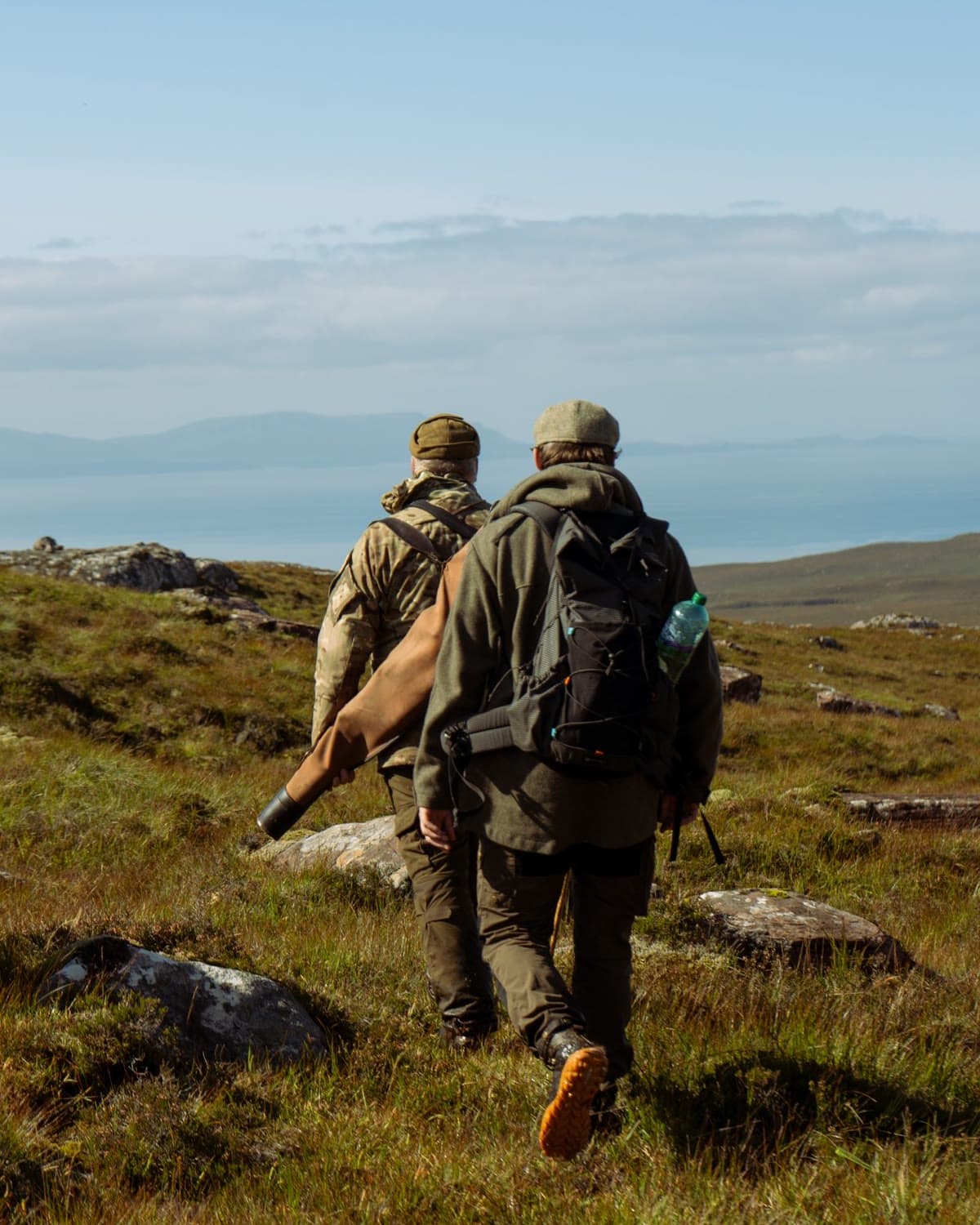 Two stalkers walk away across the hill at Diabaig towards the sea, a rifle slung in its slip