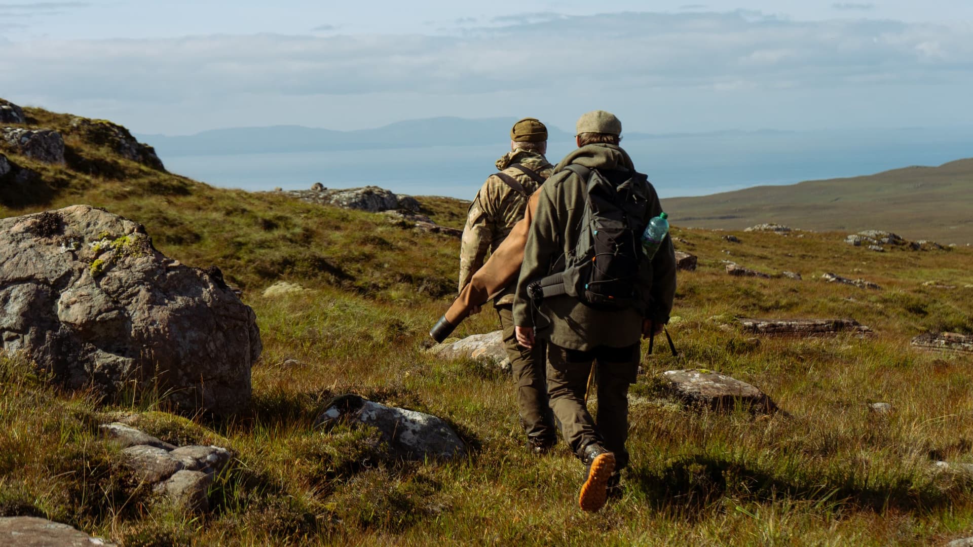 Two stalkers walk away across the hill at Diabaig towards the sea, a rifle slung in its slip