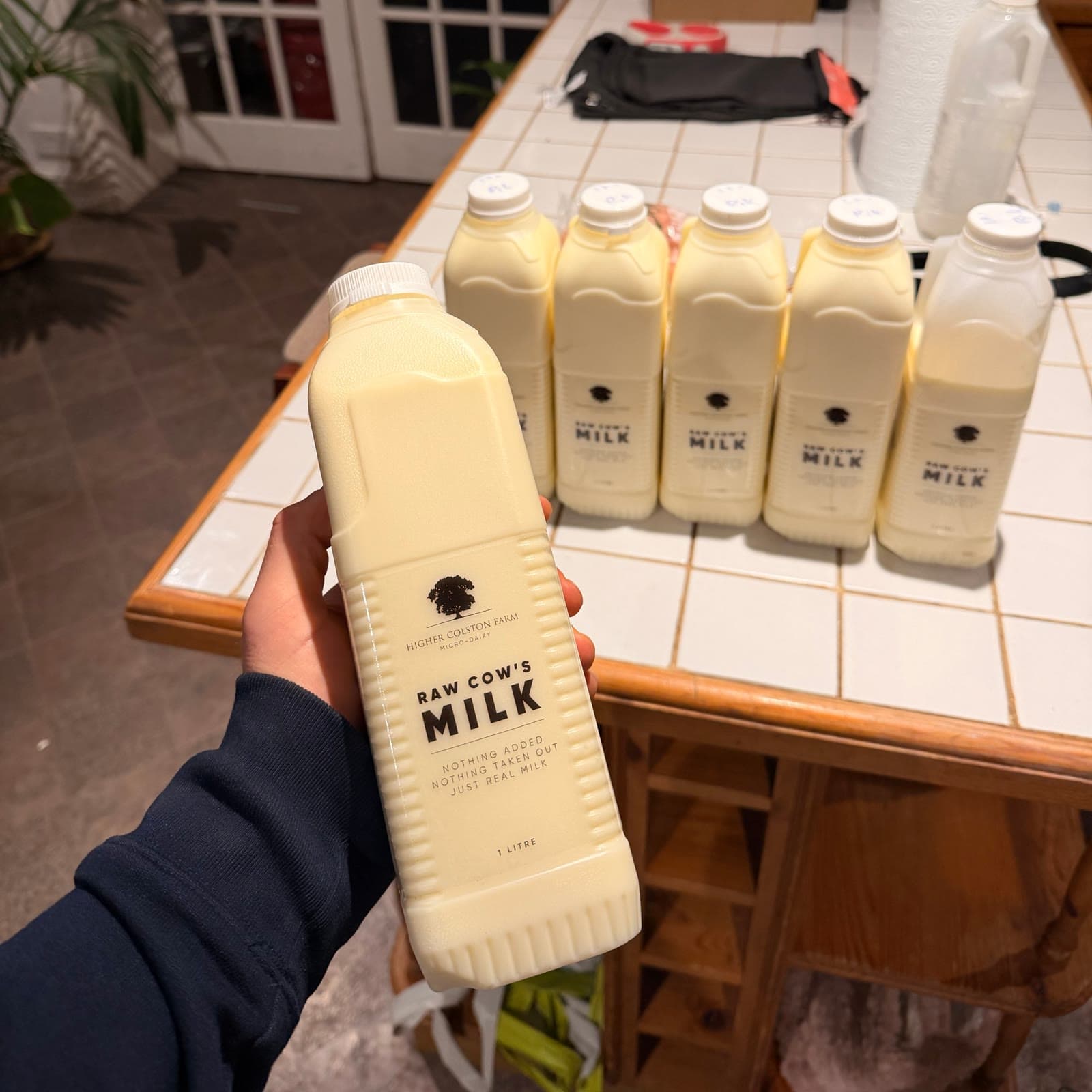 A hand holding a litre bottle of Higher Colston Farm raw cow’s milk, more bottles lined up behind