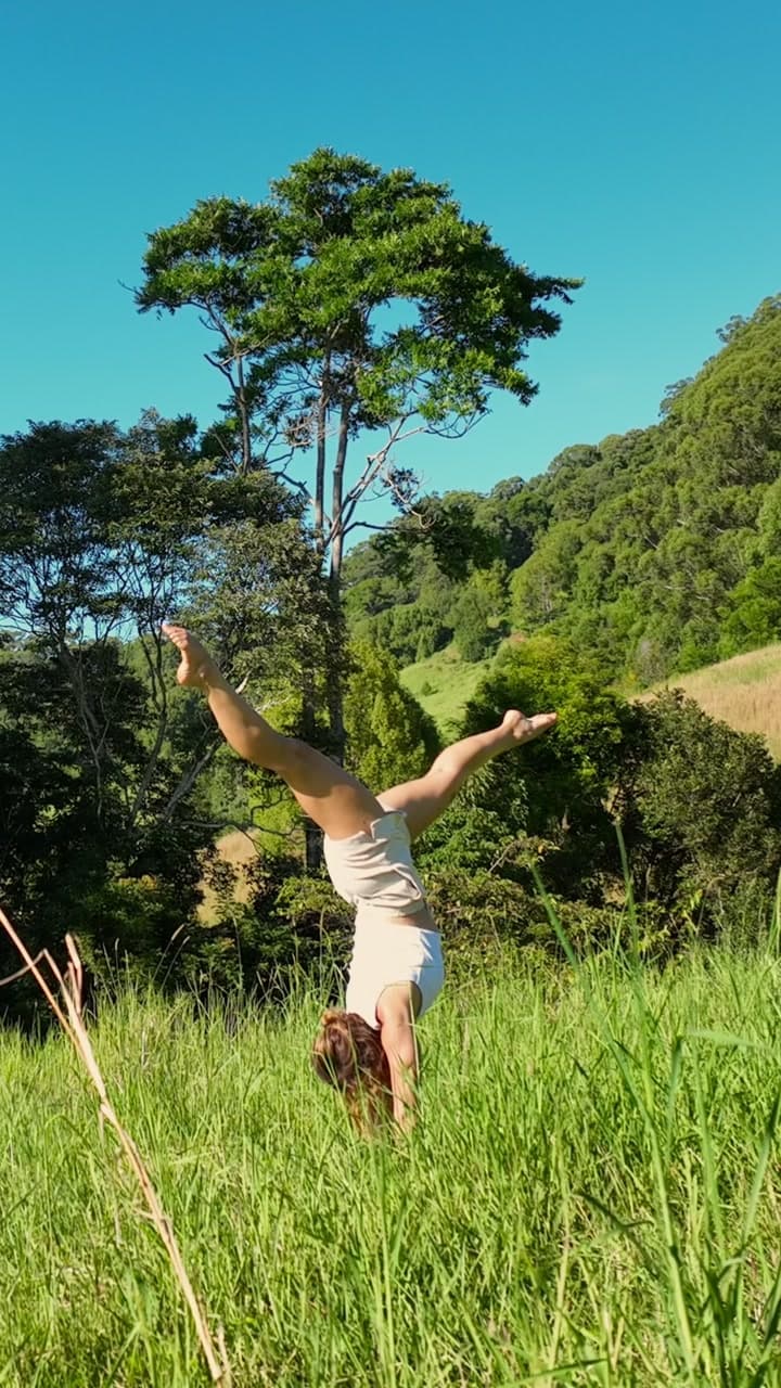A woman holding a handstand in the long grass of a sunlit meadow, wooded hills behind
