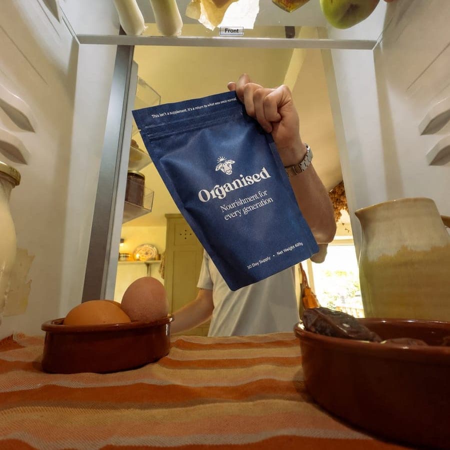 A hand lifting an Organised pouch out of the fridge, seen from inside it, past a bowl of eggs and a jug