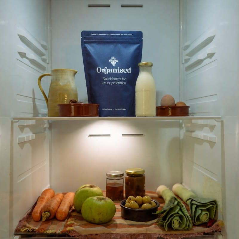 An Organised pouch on a fridge shelf between a jug and a bottle of milk, with apples, carrots and leeks on the shelf below