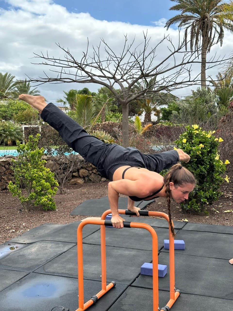 A woman holds a straddle planche on parallel bars in a garden, her body level above her hands