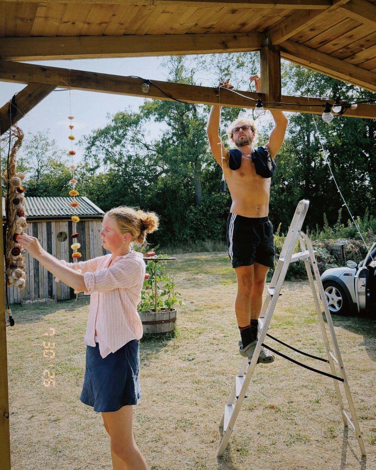 Two people hanging festoon lights and strings of garlic under a timber shelter, setting up for a gathering