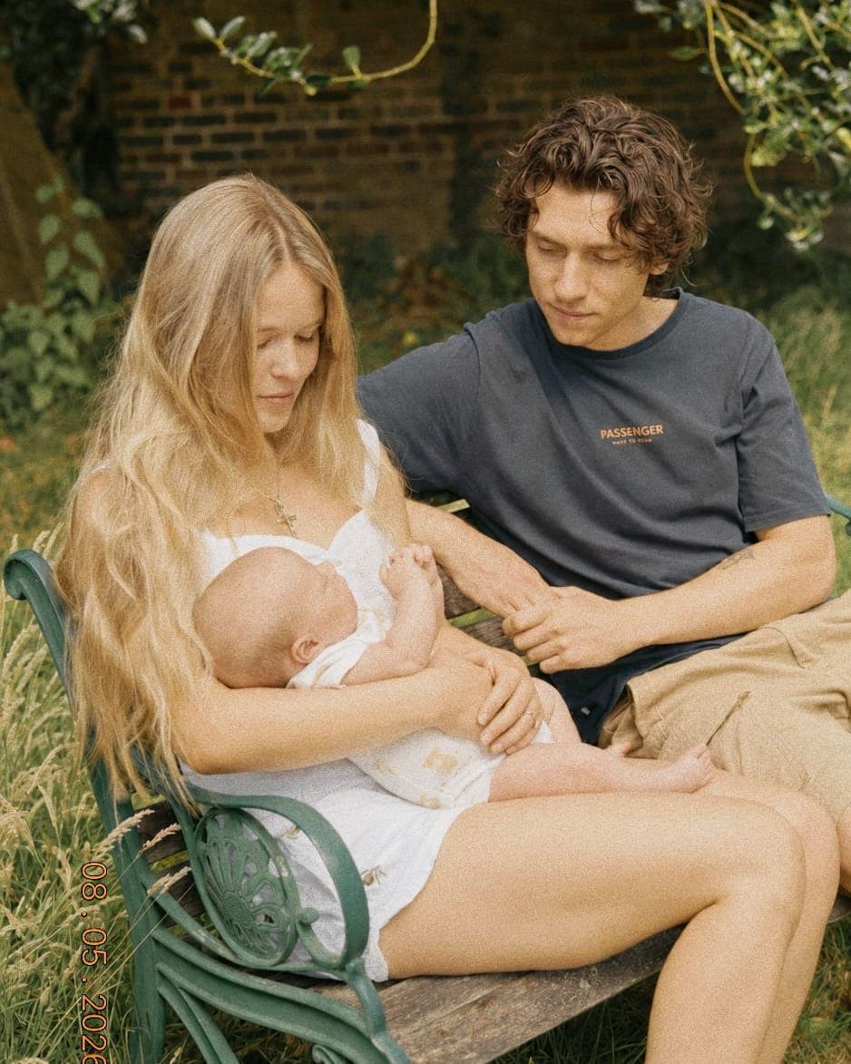 A woman with long loose hair sitting on a garden bench in summer light, a baby in her arms and a man beside her
