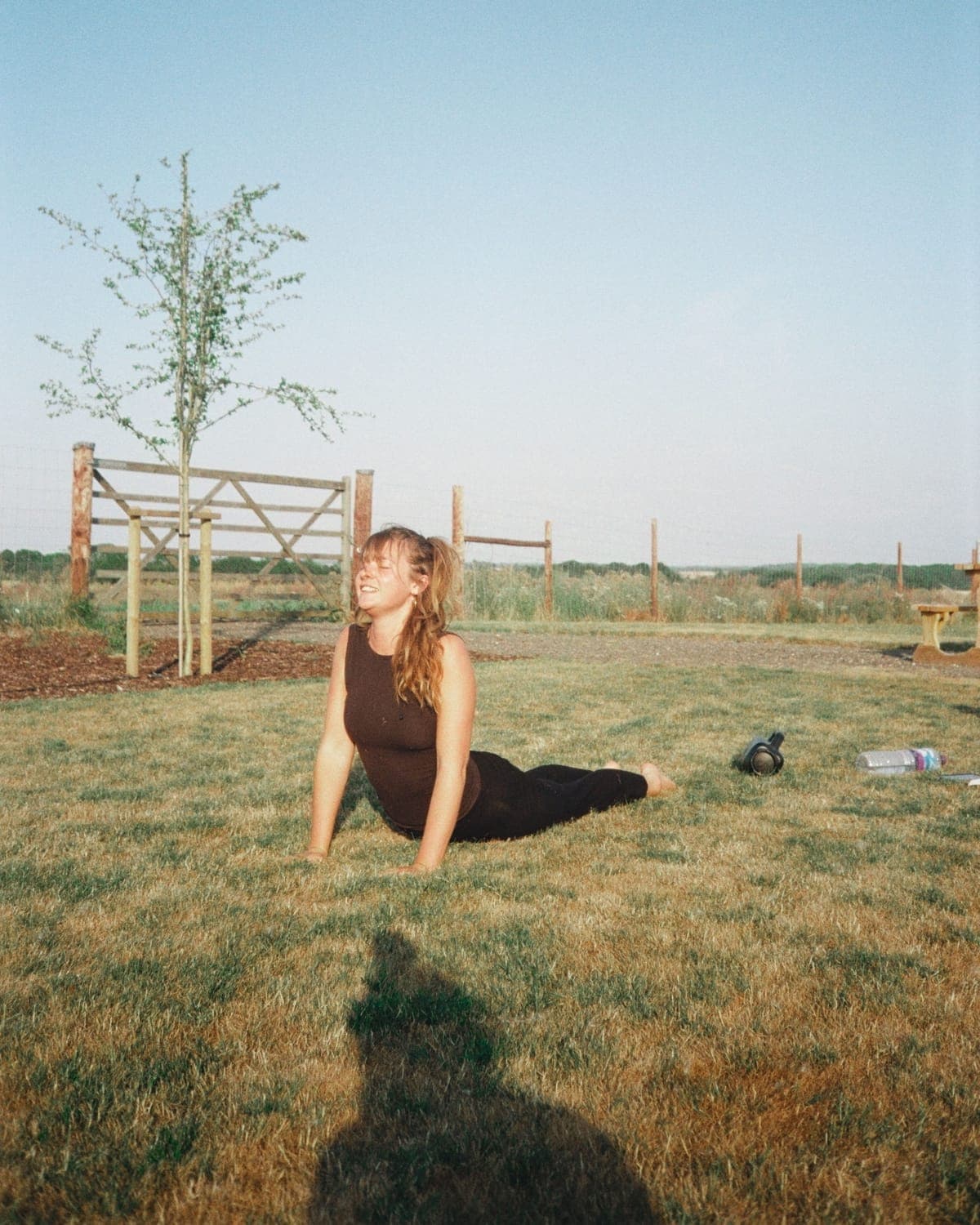 A woman stretching on mown grass in evening light, a young tree and a field gate behind her