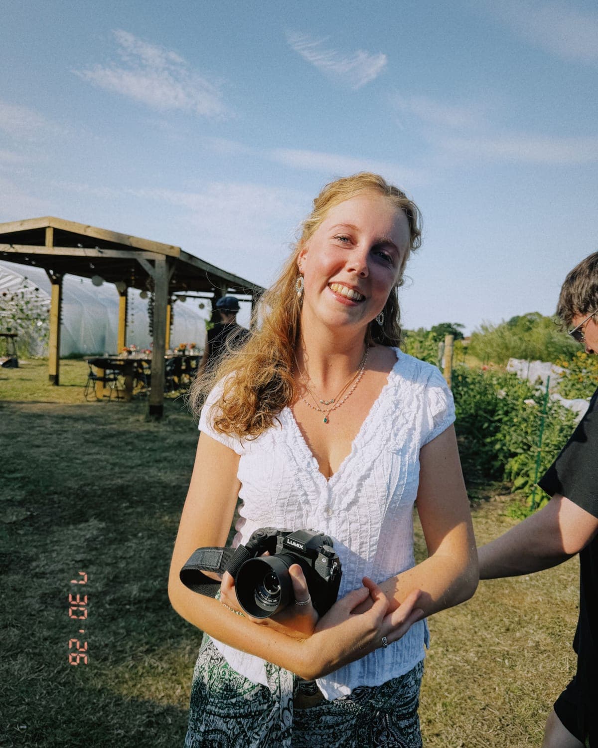 A woman smiling in full sun on a market garden, a camera in her hands, polytunnels and a long laid table behind her