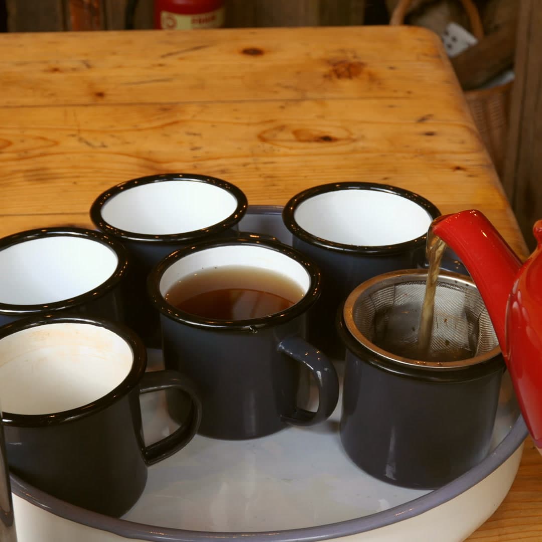 A pot pouring a warm drink through a strainer into enamel mugs on a farmhouse table