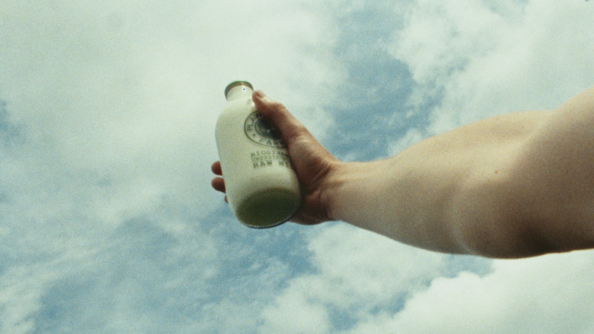A hand holds a bottle of raw milk up against a bright, cloud filled sky