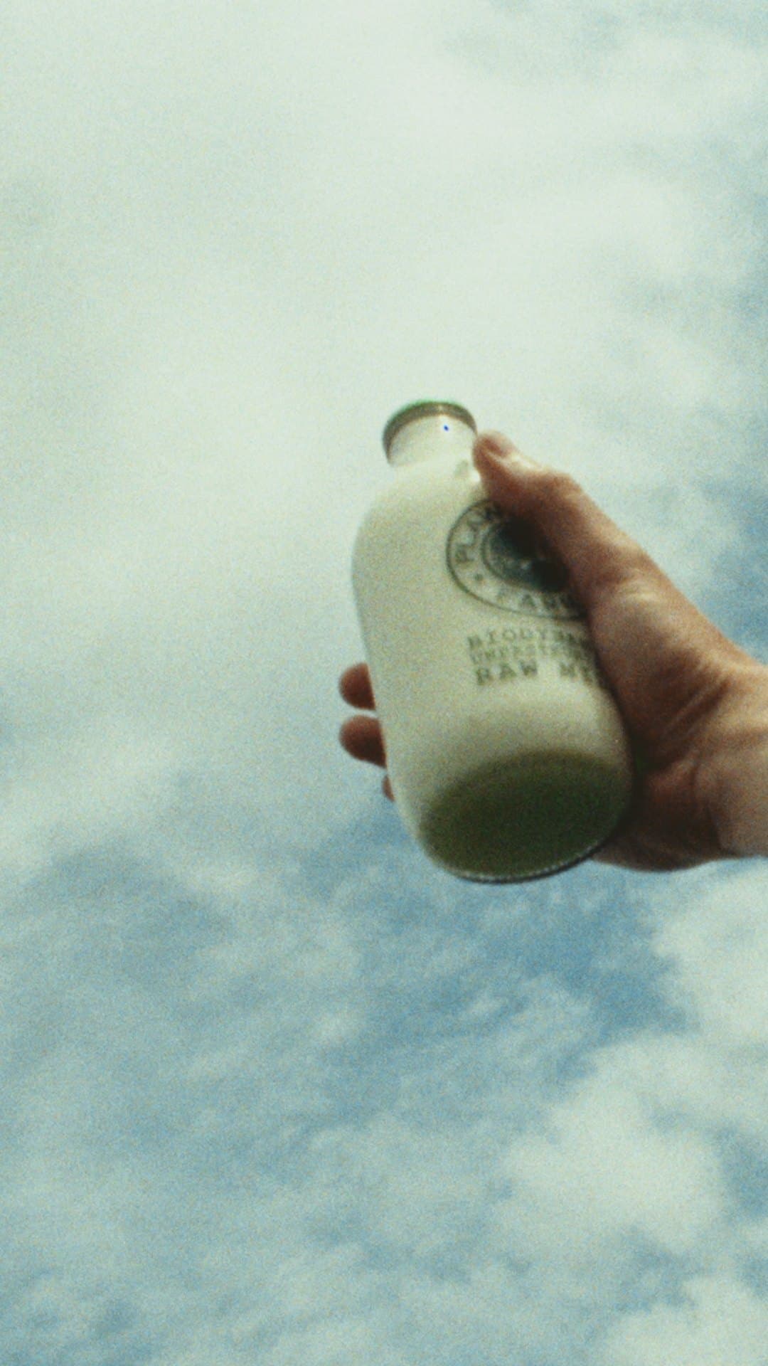 A hand holds a bottle of raw milk up against a bright, cloud filled sky