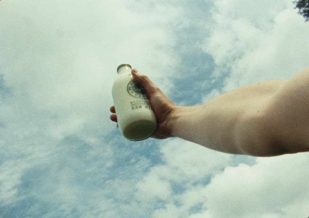 A hand holds a bottle of raw milk up against a bright, cloud filled sky