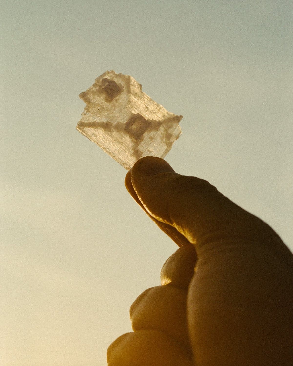 A single translucent salt crystal held between finger and thumb, backlit gold against a pale sky
