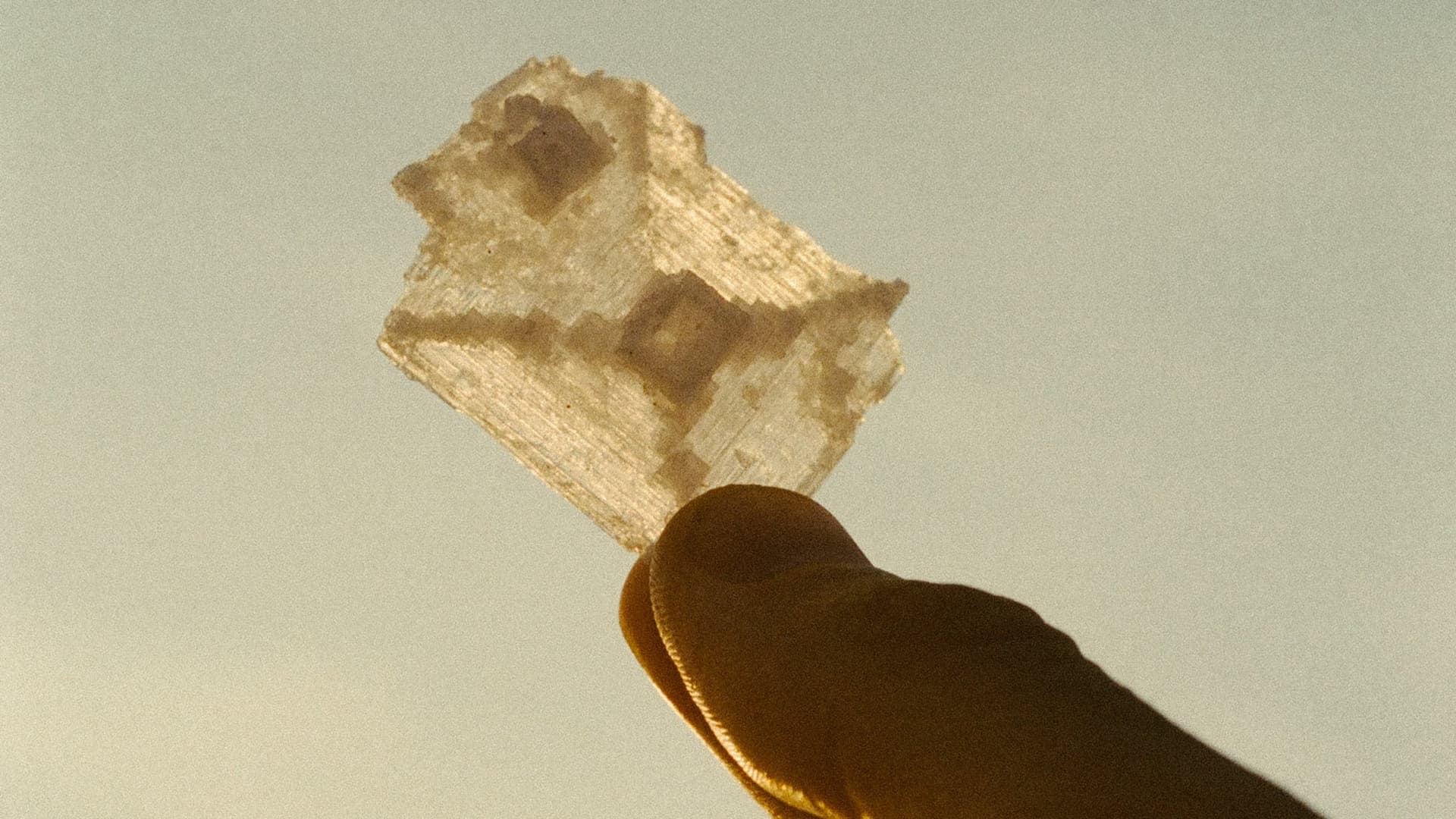 A single translucent salt crystal held between finger and thumb, backlit gold against a pale sky