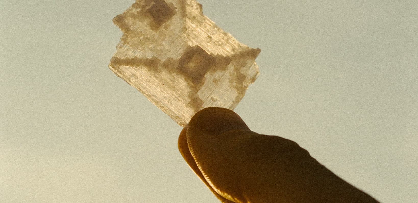 A single translucent salt crystal held between finger and thumb, backlit gold against a pale sky
