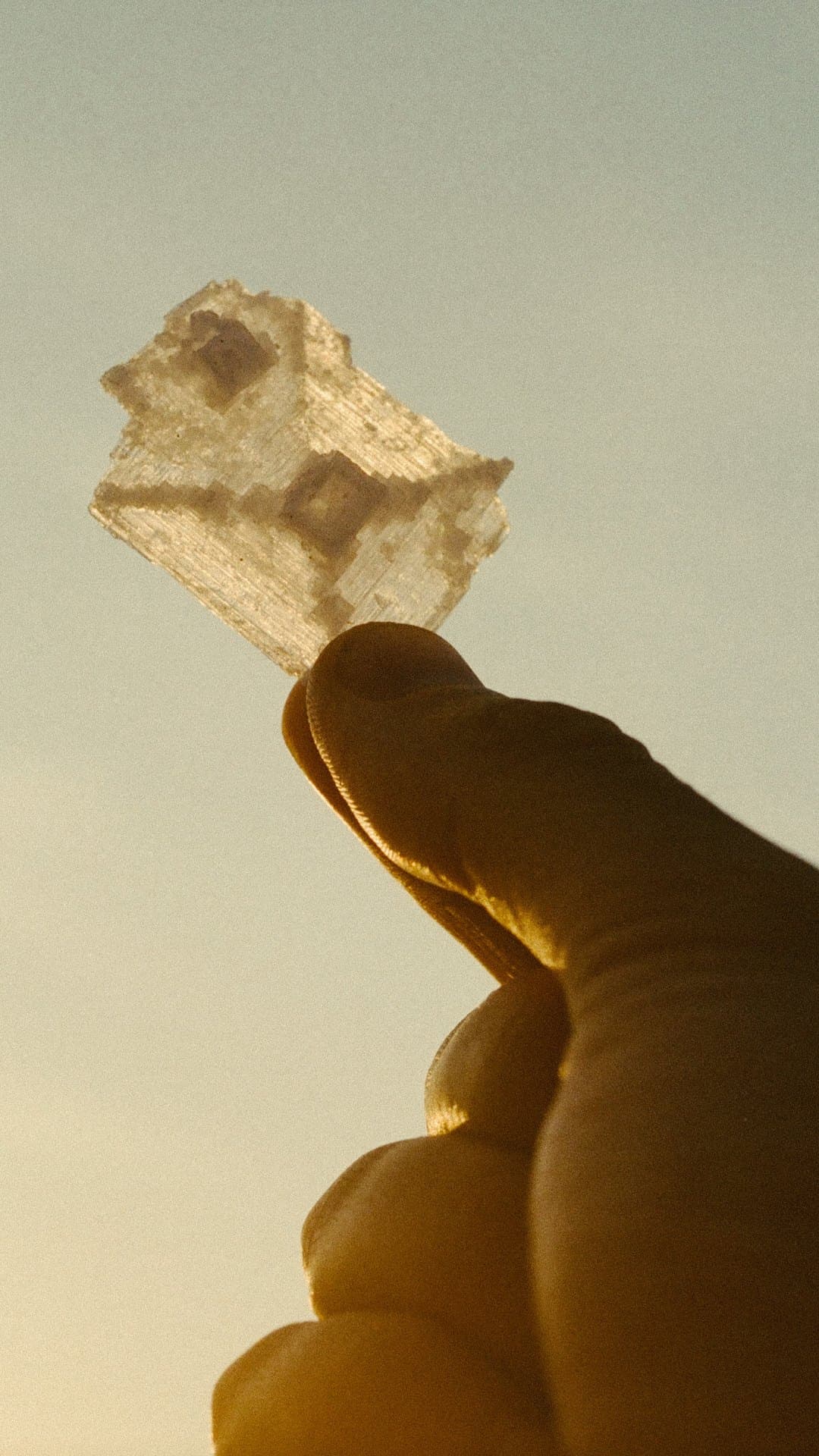 A single translucent salt crystal held between finger and thumb, backlit gold against a pale sky
