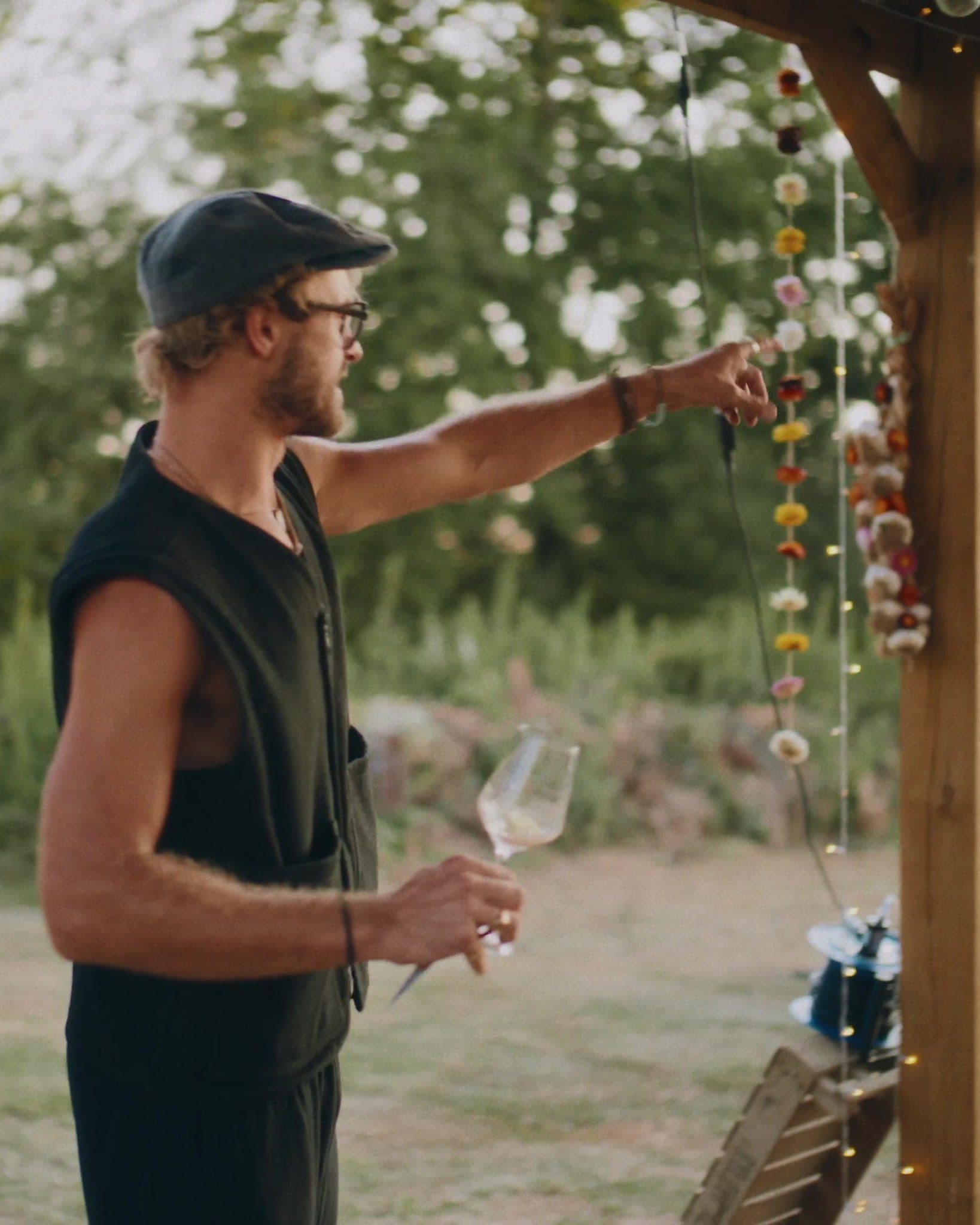 A grower in a flat cap pointing out at the growing beds in golden evening light, glass in hand