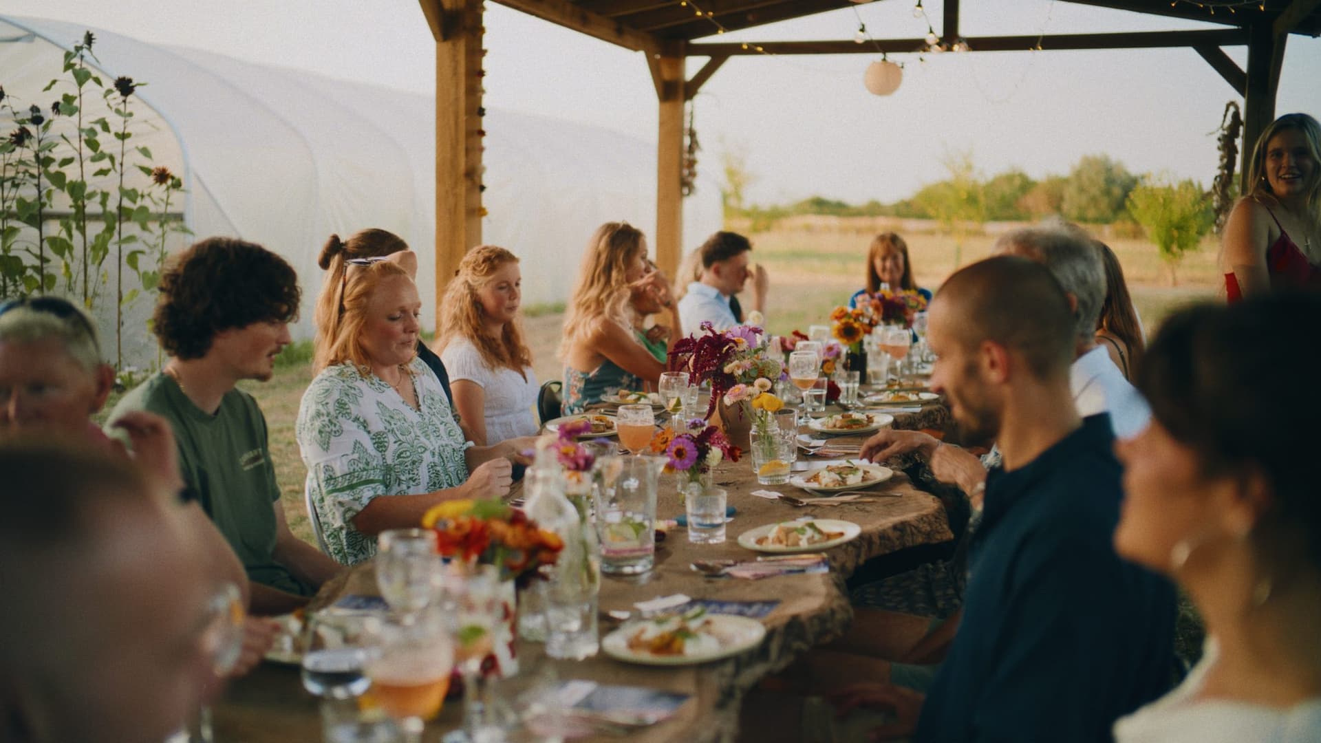 The long supper club table at golden hour, guests seated under the timber canopy with the fields behind