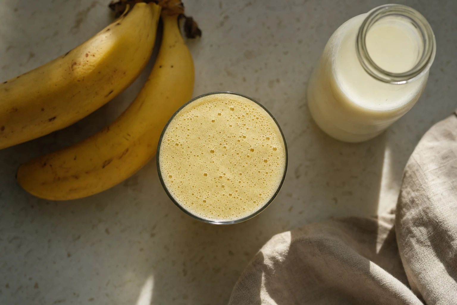 A pale-yellow shake in a clear glass, with a banana and bottle of raw milk beside it
